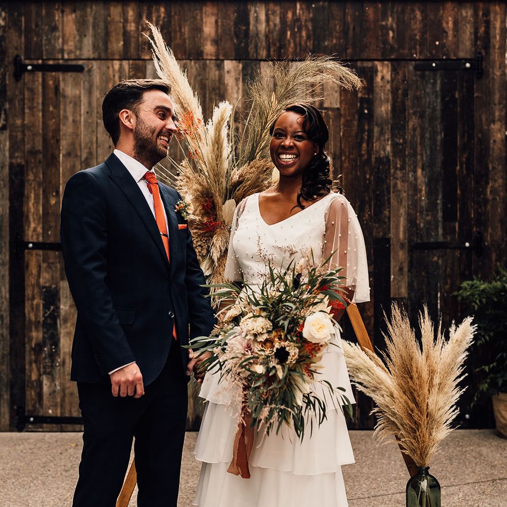 Black bride in bridal separates and groom in a navy suit standing at their wooden frame altar decorated with pampas grass and giant bottles 