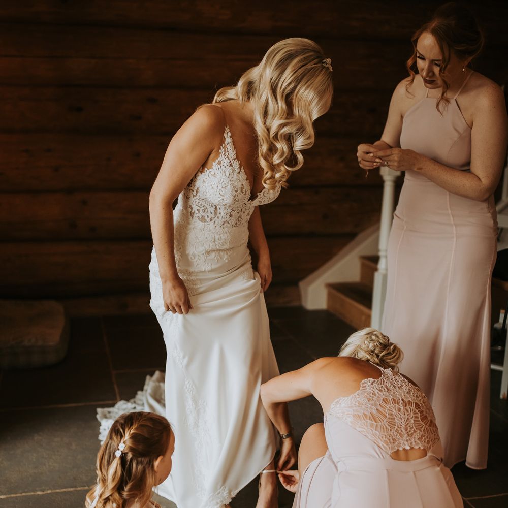 Bride with curled blonde hair and lace wedding dress stands whilst bridesmaids in pink bridesmaid dresses help her put on her shoes before wedding
