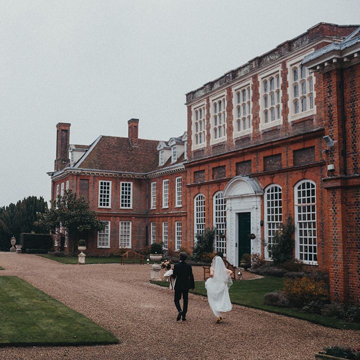 Bride & groom walk in front of Gosfield Hall on their wedding day 