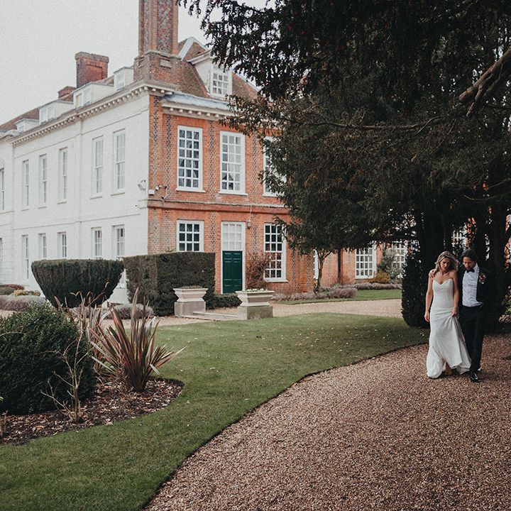 Bride & groom walk across gravelled pathway in front of Gosfield Hall 