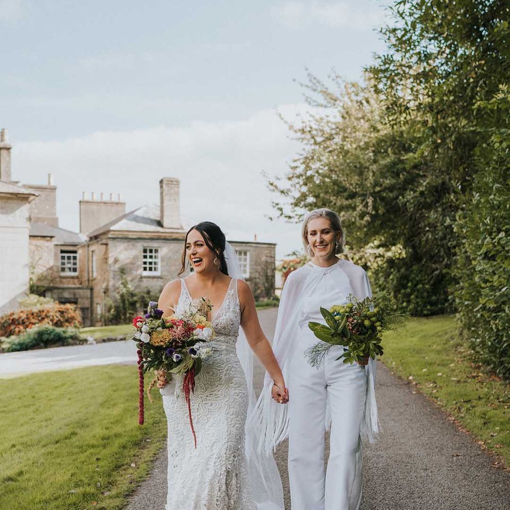 Lesbian wedding with two brides in a lace wedding dress and bridal jumpsuit and cape holding a brightly coloured wedding bouquet 