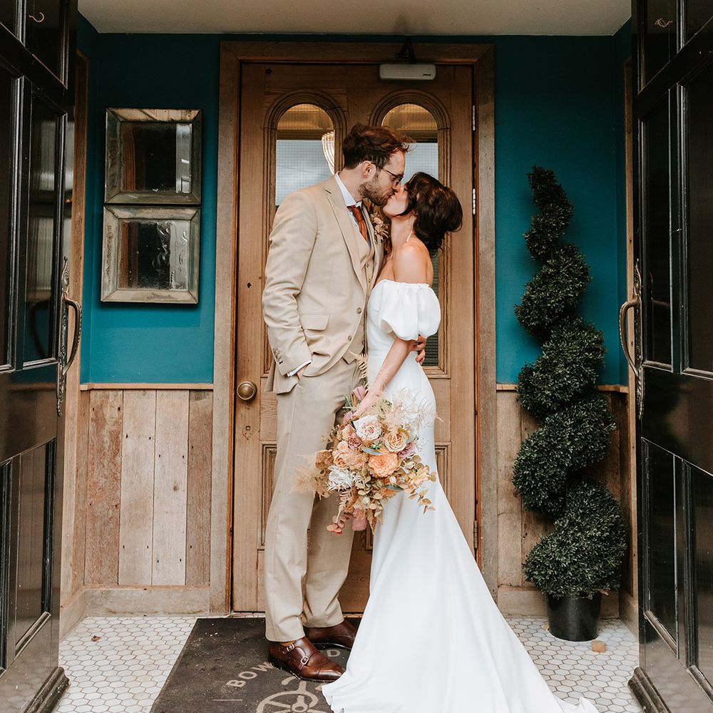 Groom in three piece beige suit kisses bride outside wedding venue with bride in puff sleeve sweetheart wedding dress 