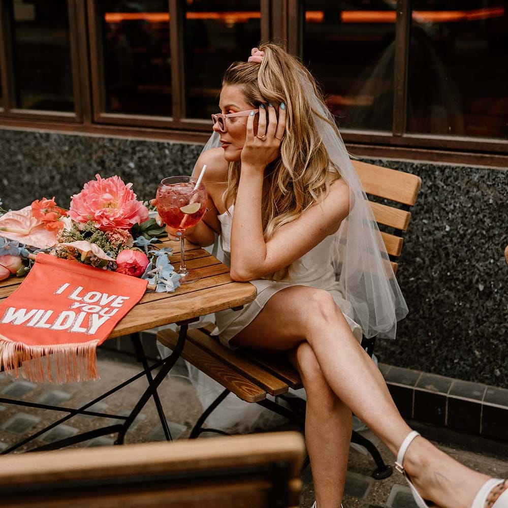 Bride in a retro pink sunglasses and short wedding dress drinking fruity cocktail with a fabric wedding banner on the table 