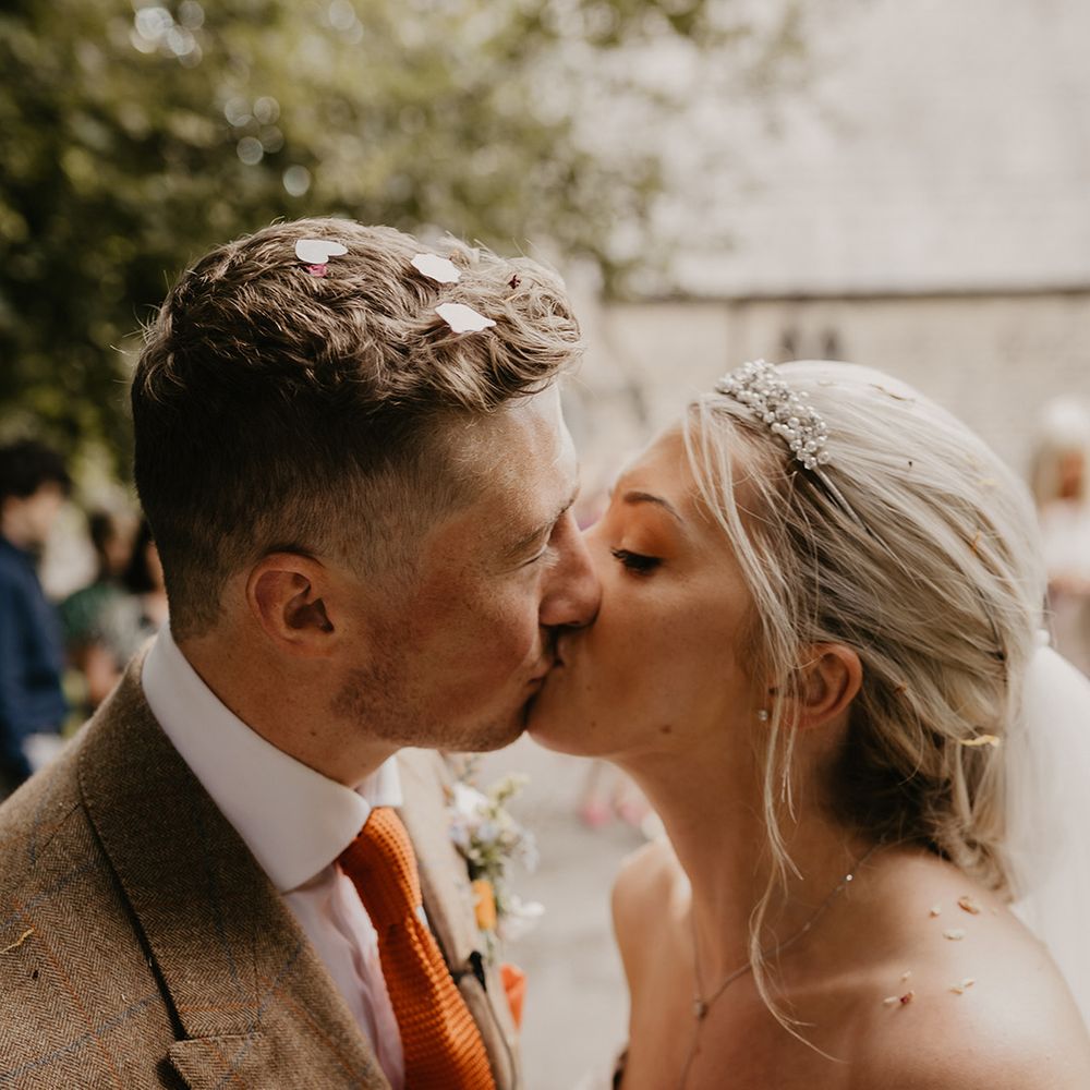 Bride in pearl tiara kisses the groom in brown suit and orange tie