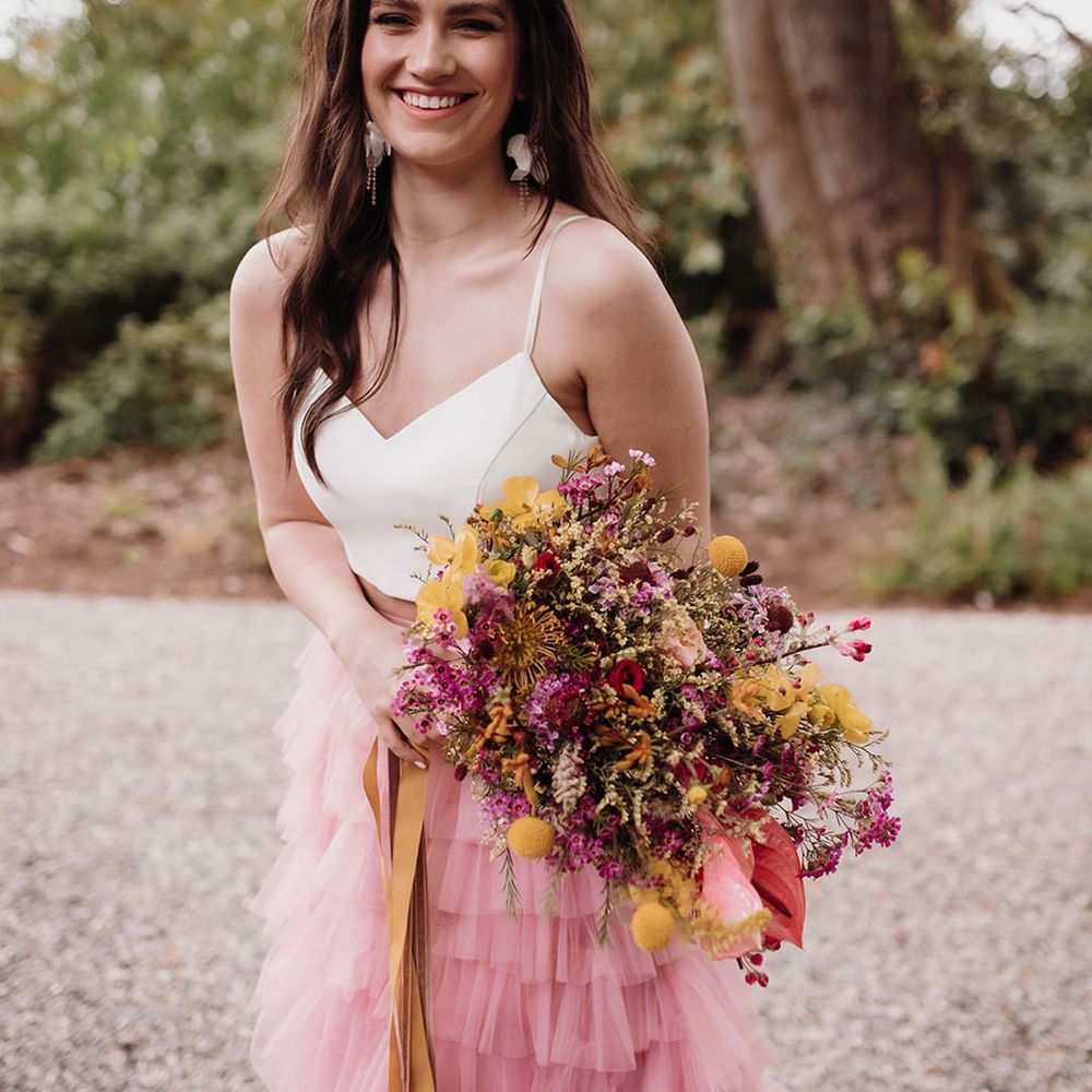 Bride with long wavy hair in a white top and pink skirt holding a pink and yellow wildflower wedding bouquet 