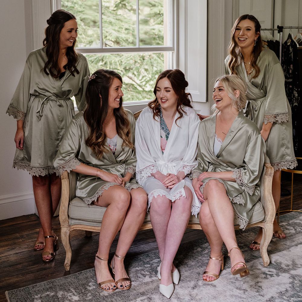 Bride in white satin robe getting ready with bridesmaids in green satin robes and matching gold sandals for wedding day 