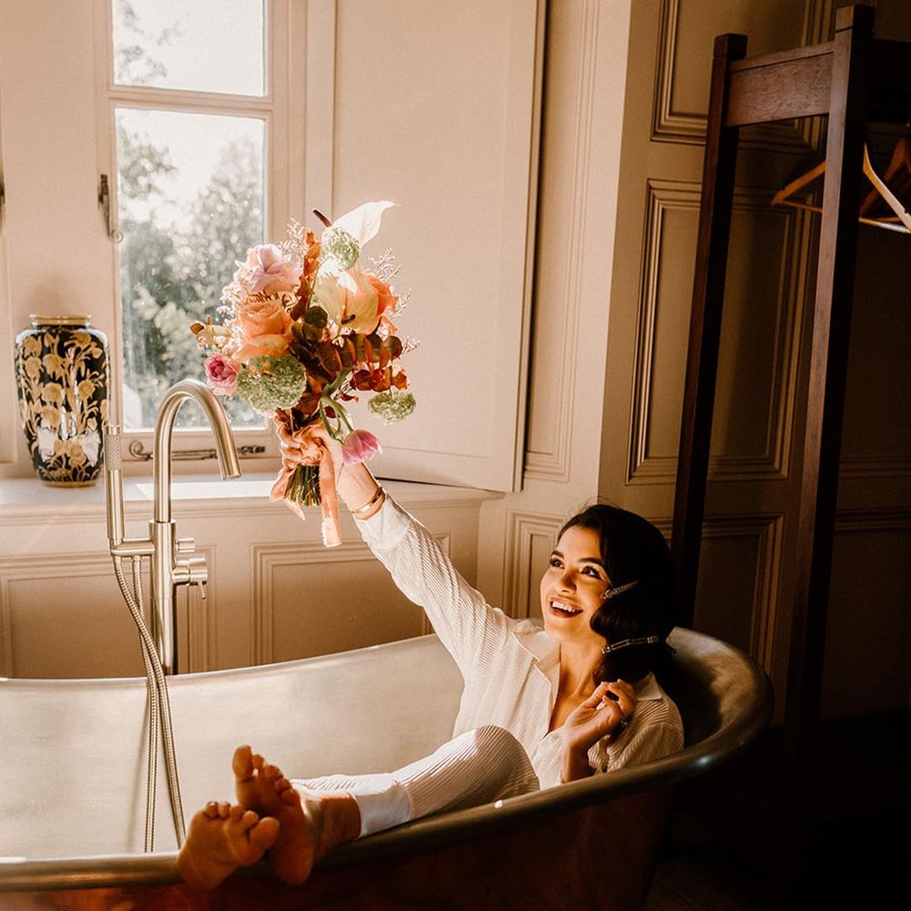 Bride in white bridal pyjamas in a copper bath tub holding her autumnal wedding bouquet 