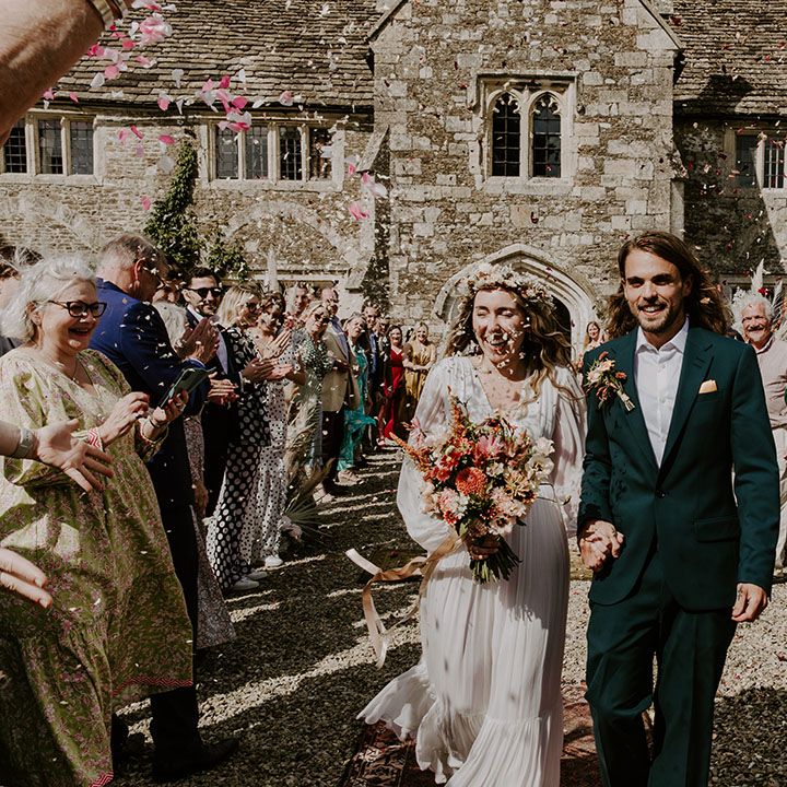 Groom wears green suit with floral buttonhole and pocket square and walks with bride carrying floral bouquet