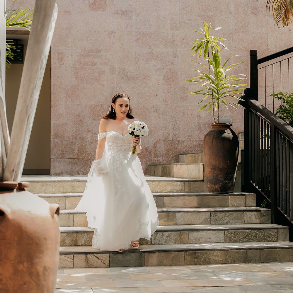 Bride in off the shoulder lace wedding dress with sweetheart neckline and lace floral details and cathedral length veil holding white tropical bouquet 