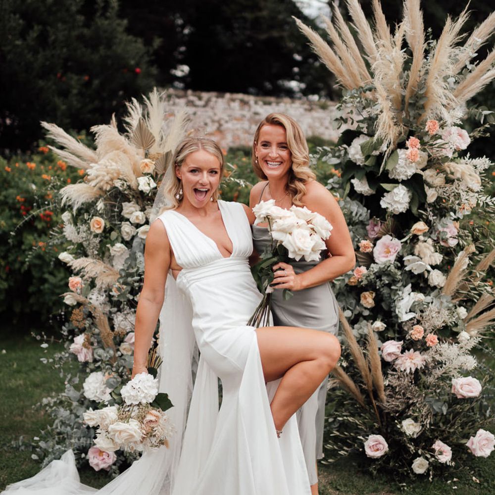 The bride and bridesmaid pose together in front of the flower columns with pink roses and pampas grass 