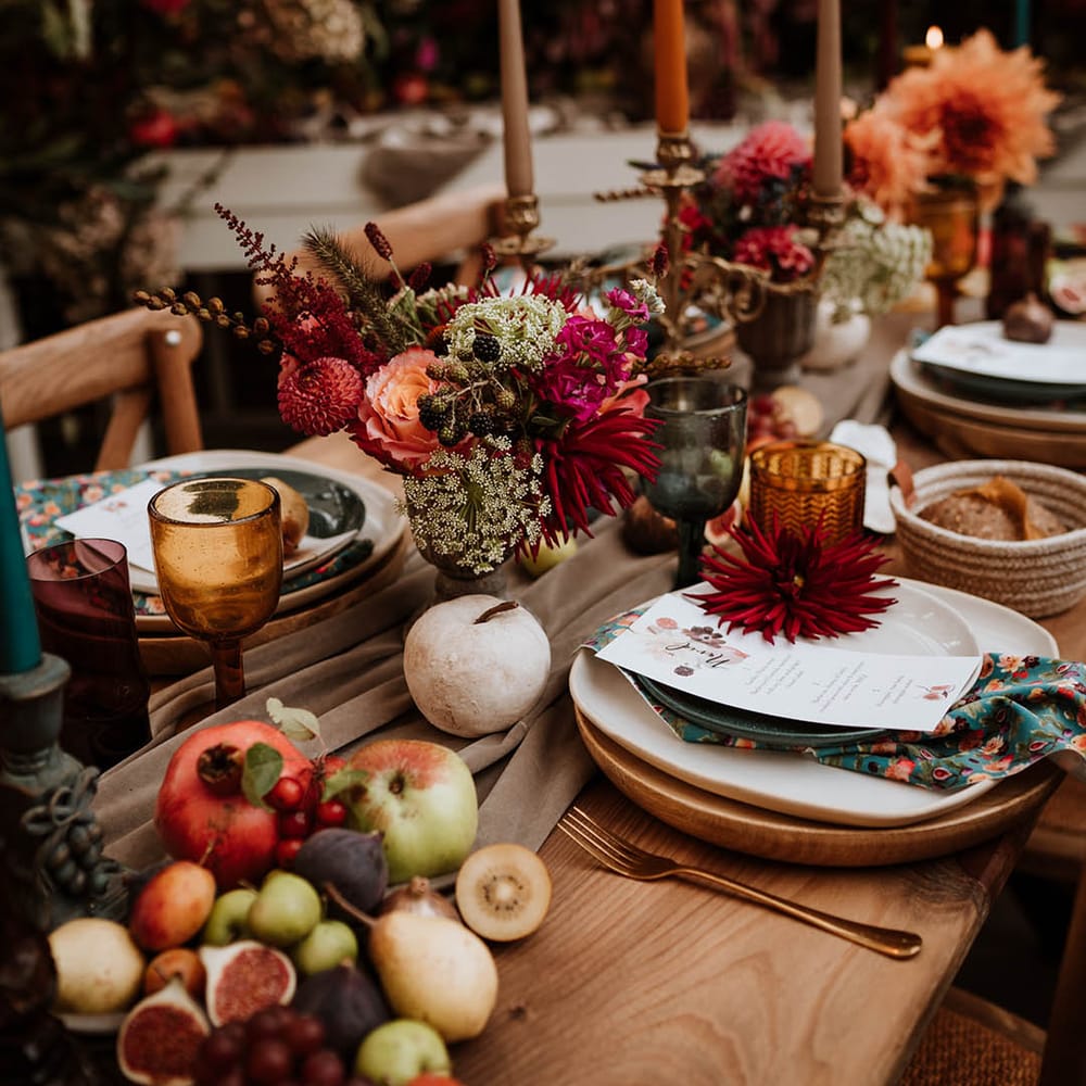 Neutral toned tablerunner on rustic wooden table, orange, green and red coloured glasses, earthenware, seasonal fruit and vegetables, red, brown and green tapered candles in gold candlesticks and clear vases with autumnal coloured flowers