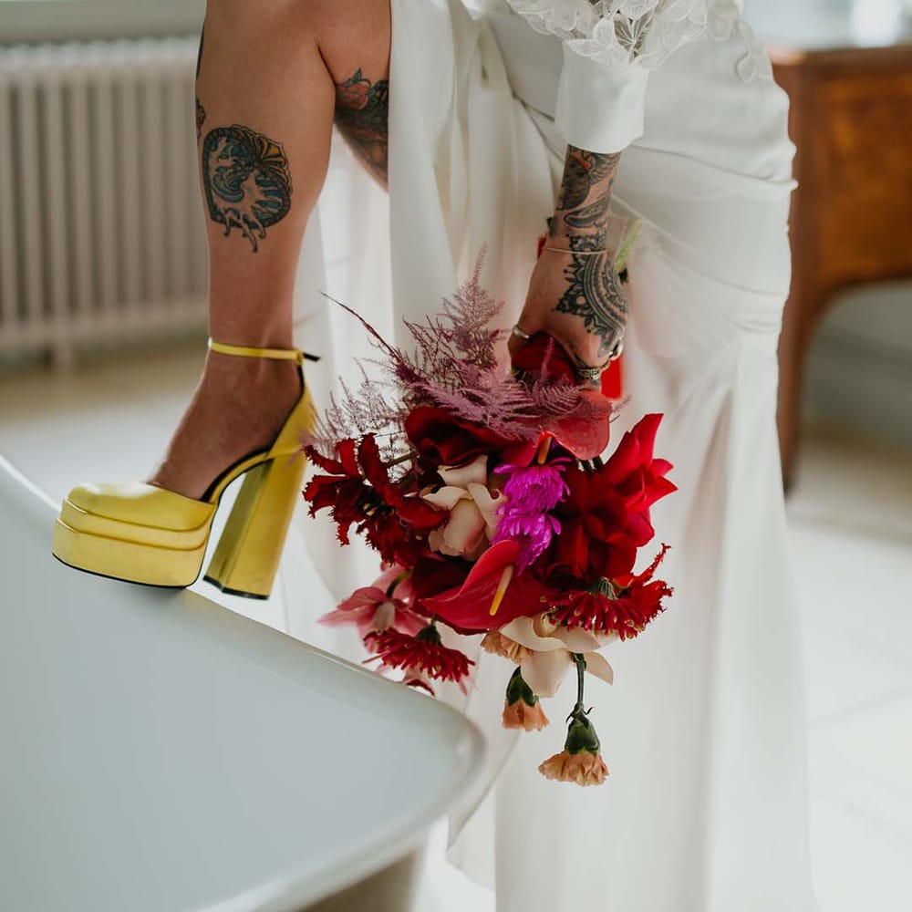 Bride in two piece bridal separates and yellow chunky platform heels holding bridal bouquet with magenta and hot pink dahlias, orchids, roses, amaryllis, anthuriums and dried flowers 