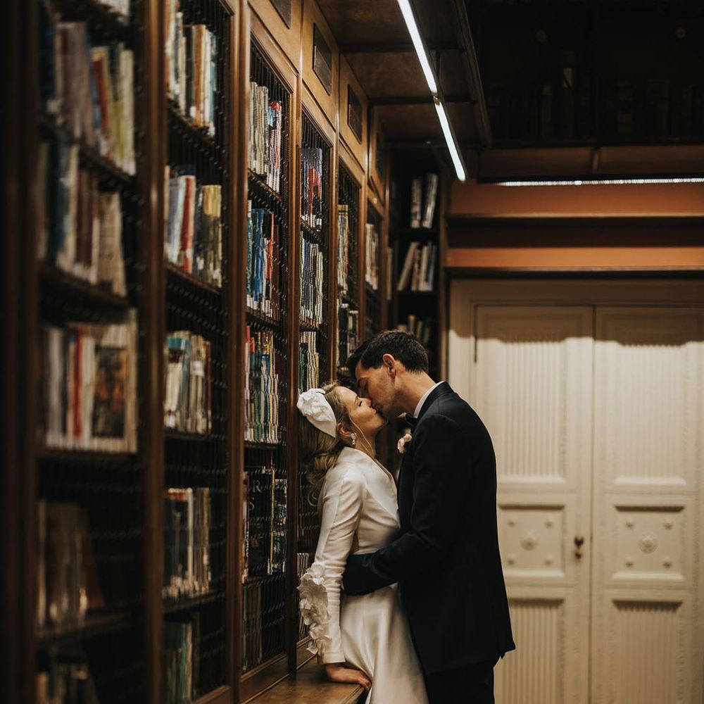 The groom kisses the bride against the bookcase for a stolen moment at their Bridgerton inspired wedding 