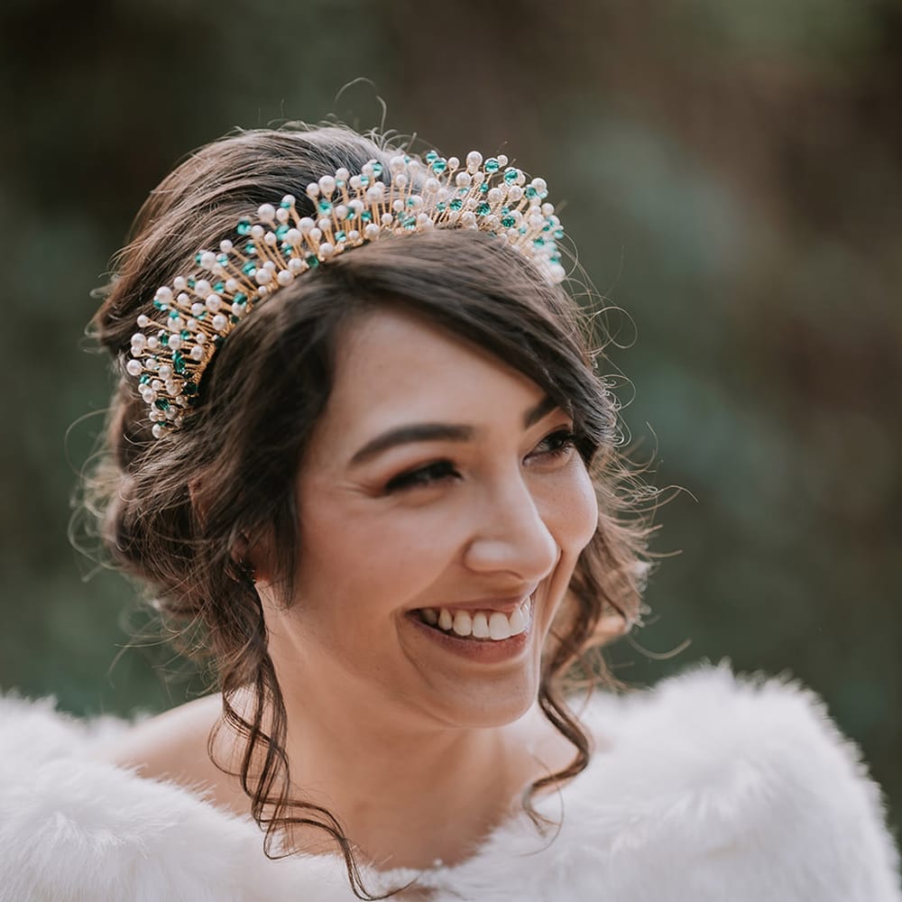 Bride in cropped faux fur bridal cover up and gold, pearl and emerald bridal crown smiling