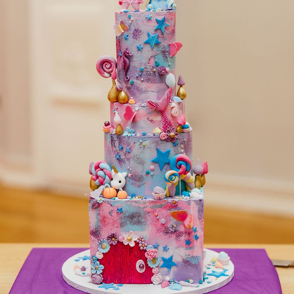 Four tier pink and purple iced wedding cake with cartoon and fairy decorations, lollipop, star, and flower icing and personalised pink and blue wedding cake toppers 