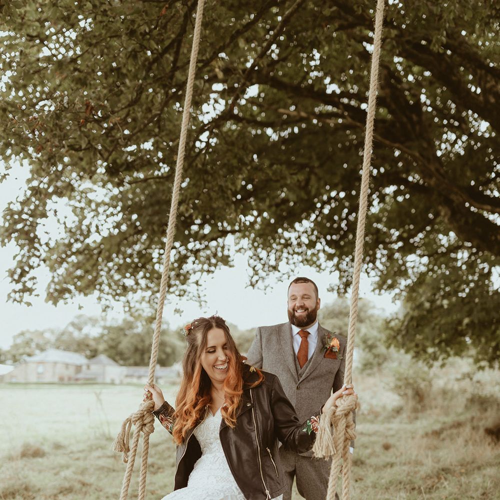 Groom in there piece tweed suit with autumnal orange tie pushing the bride on a big swing wearing a personalised leather jacket and fitted lace wedding dress 