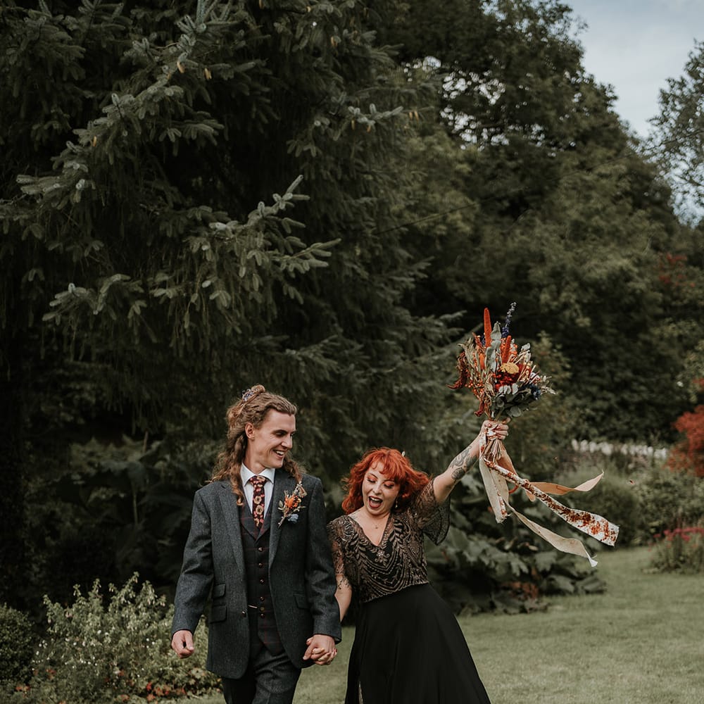 Groom in dark grey three piece suit and burgundy flower patterned tie and bride in black dress for wedding in Devon 