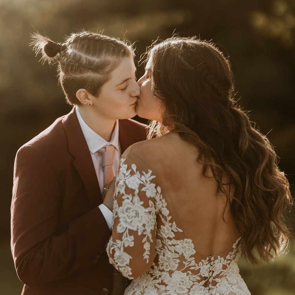 Bride in maroon bridal suit with grey waistcoat, dusky pink tie and autumnal dried flower boutonniere kissing bride in long sleeve lace illusion sleeve wedding dress with overlay at golden hour wedding photoshoot