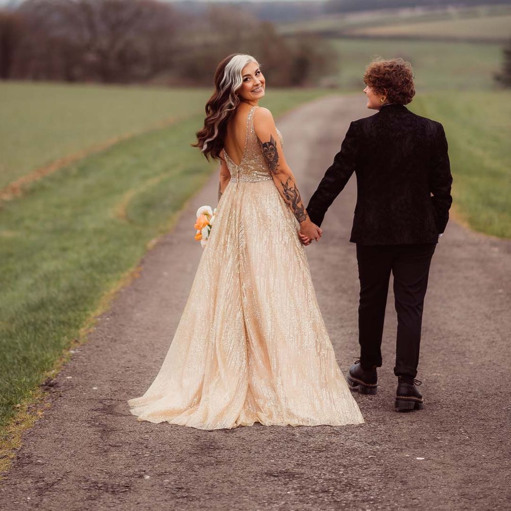 Bride looking back at camera in sleeveless deep v neck sparkly gold wedding dress holding up orange garden rose, white poppy, light pink carnation and foliage bridal bouquet wrapped in sparkly gold ribbon with bride in dark bridal suit at Kingscote Barn Tetbury 