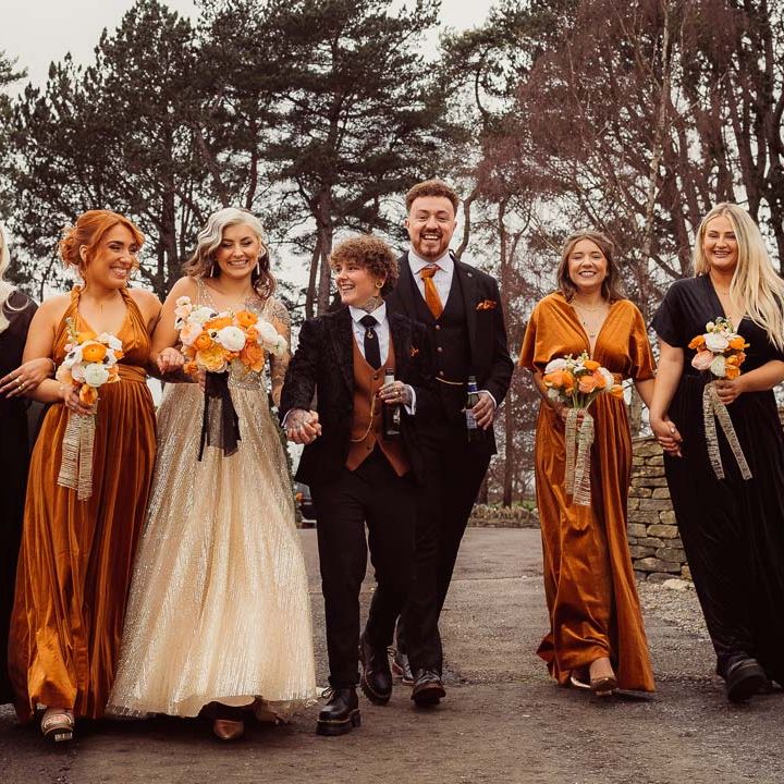 Bride in dark velvet bridal jacket with rust waistcoat and pocket square, black suit trousers, platform Dr Martens and gold pocket watch walking hand in hand with bride in sleeveless deep v neck sparkly gold wedding dress with sheer and gold polka dot wedding veil holding orange garden rose, white poppy, light pink carnation and foliage bridal bouquet wrapped in sparkly gold ribbon with bridesmaids in black and orange mismatched bridesmaid dresses and groomsmen in dark three piece suit with orange tie and pocket square 