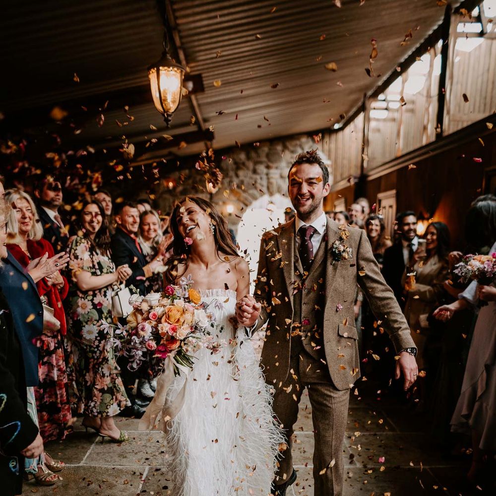 Bride in strapless ivory fringed crepe gown and chapel length veil holding mixed bridal bouquet with garden roses, carnations, peonies and foliage doing colourful dried petal wedding confetti exit with groom in chocolate brown grooms suit at Willow Marsh Farm Loughborough 