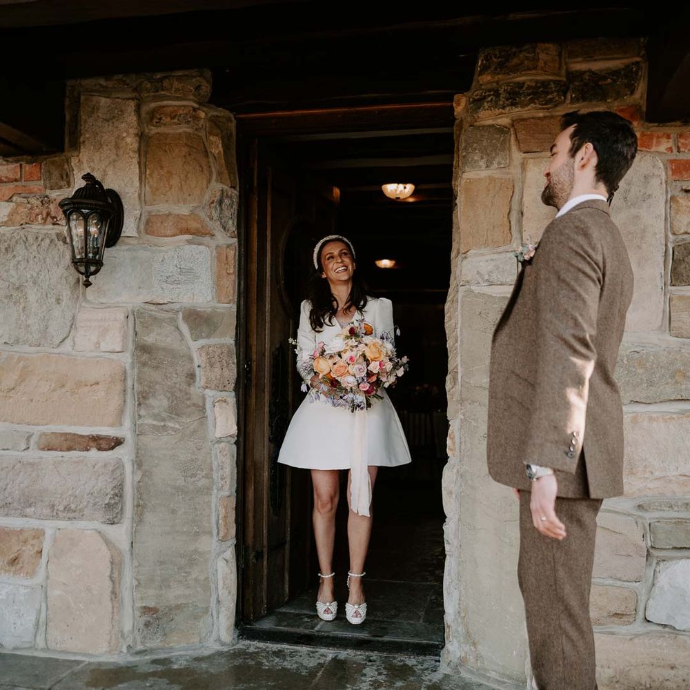Bride in long sleeve short wedding dress, pearl bridal headband and platform pearl wedding heels holding mixed bridal bouquet with garden roses, carnations, peonies and foliage looking at groom in brown grooms suit 