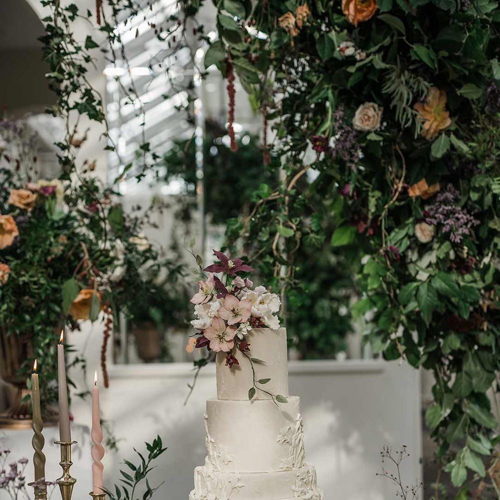 Four tier white wedding cake with intricate icing details and dried flower wedding cake topper on wooden table with twisted tapered candles in neutral shades in gold candlesticks surrounded by wildflower and foliage wedding flower arrangements