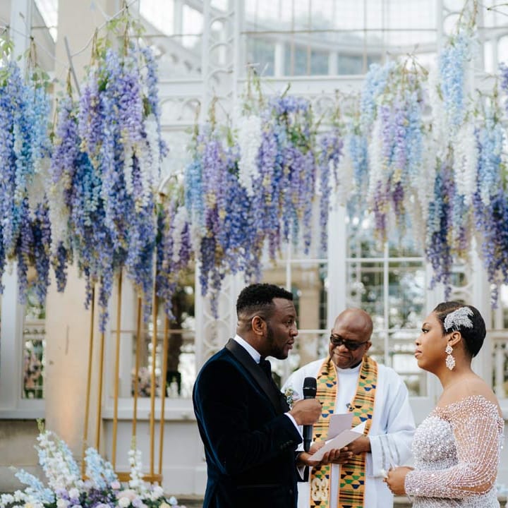 Bride in illusion v neck sheer off shoulder long sleeve pearl embellished wedding gown standing with groom, reading his vow renewal, wearing velvet midnight blue double breasted groom’s blazer, velvet bowtie and peach garden rose boutonniere underneath suspended pastel flower arrangements at Syon Park
