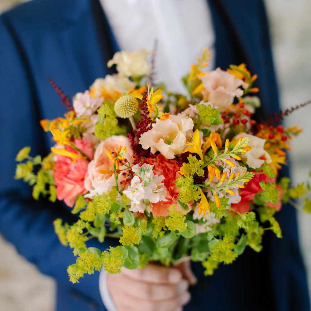 The groom holding a bouquet of green and pink wildflowers, including carnations and red berries