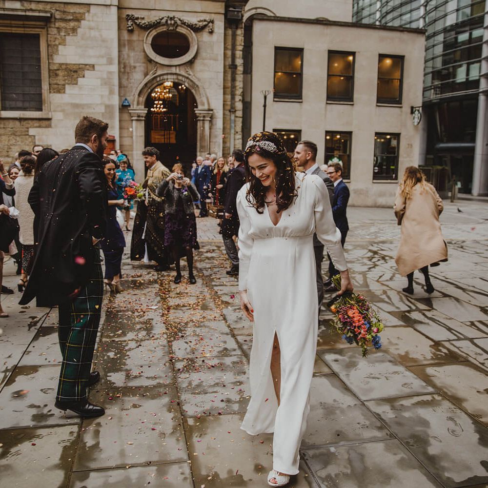 Bride wearing long sleeved wedding dress with split front. In the background the groom is celebrating with guests outside their London wedding venue