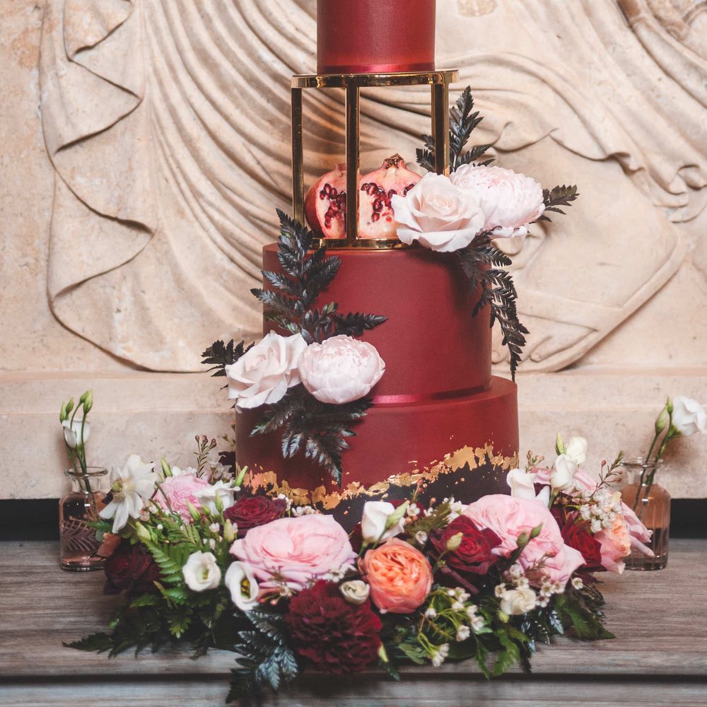 Luxury burgundy wedding cake with elevated third tier on a gold stand decorated with red, white and pink flowers, pomegranates and foliage.