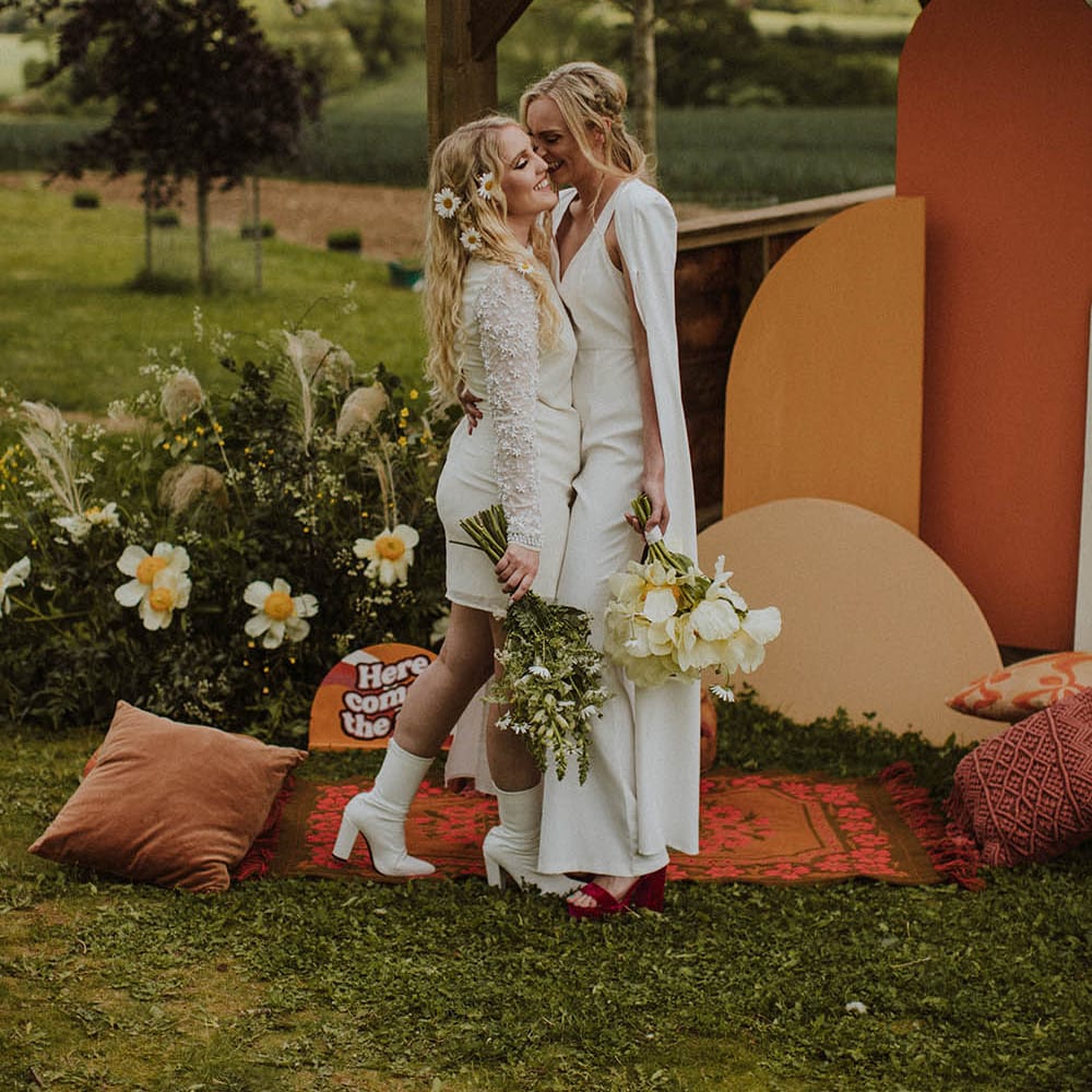 Bride in a jumpsuit with cape embracing her bride in a short wedding dress and bridal boots at an outdoor sixties wedding 