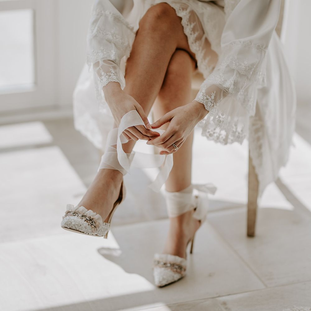 Bride in a white getting ready robe putting on her Diane Hassall wedding shoes
