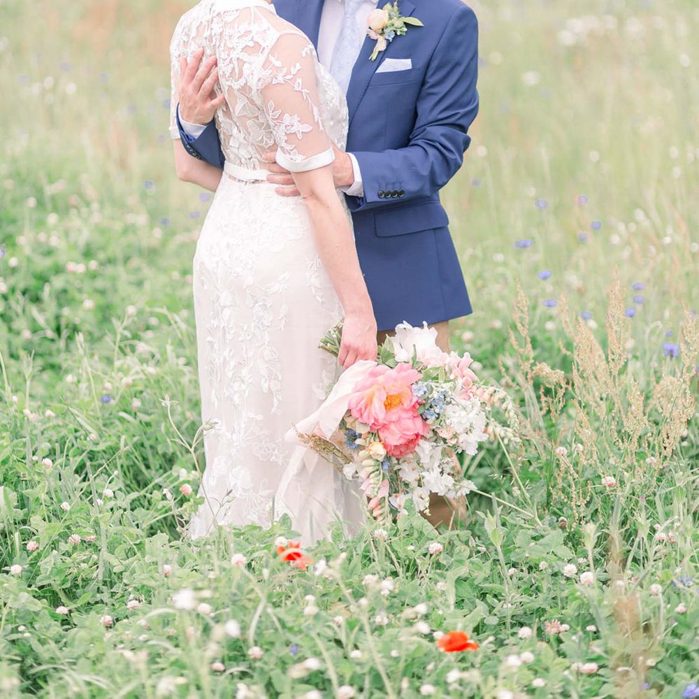Groom in navy blazer embracing his bride in a lace wedding dress in a field 