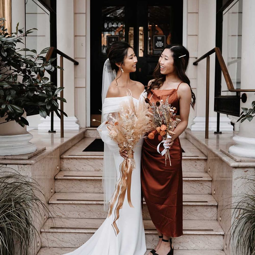 Bridesmaid in burnt orange silky dress smiling with bride in long white wedding dress both holding dried bouquets full of a colour palette of autumn colours 