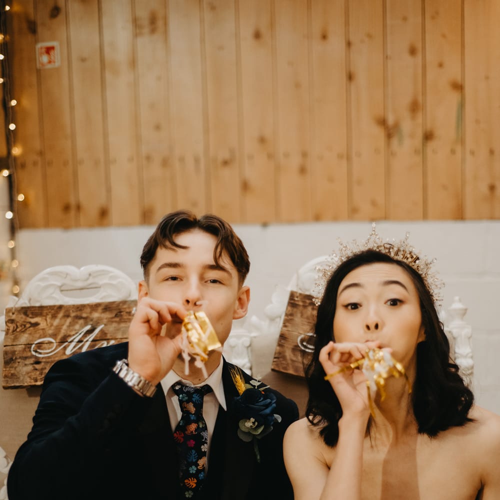 Groom in blue suit and floral tie and bride in strapless wedding dress and headpiece blow party blowers during rustic wedding ceremony in Lancashire