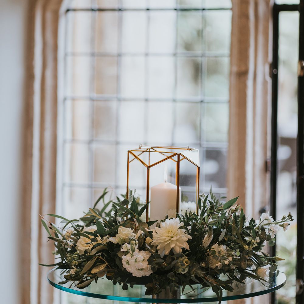 Candle in glass candle holder surrounded by wreath of white and green flowers on glass table at Notley Abbey wedding