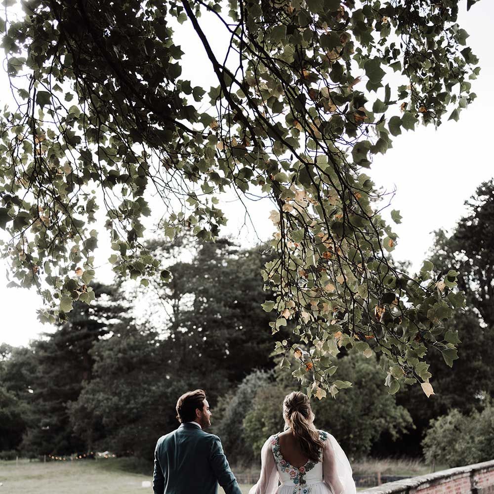 Groom in blue suit walks holding hands with bride in low back embroidered Victoria Sanders wedding dress with long sleeves through the grounds of Iscoyd Park