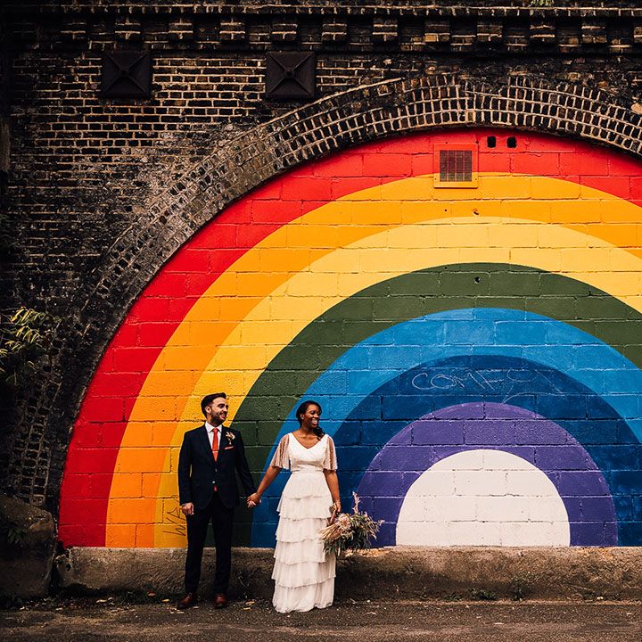 Black bride in Story Of My Dress bridal separates and groom in a navy suit standing in front of a giant street art rainbow motif in Brixton 