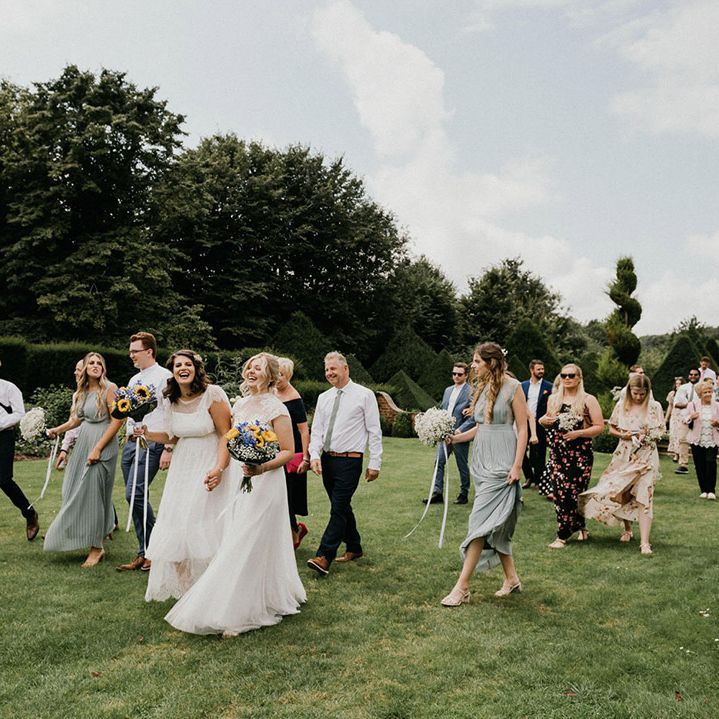 Blonde and brunette brides laugh together as their wedding party walks with them around the grounds of the barn wedding venue