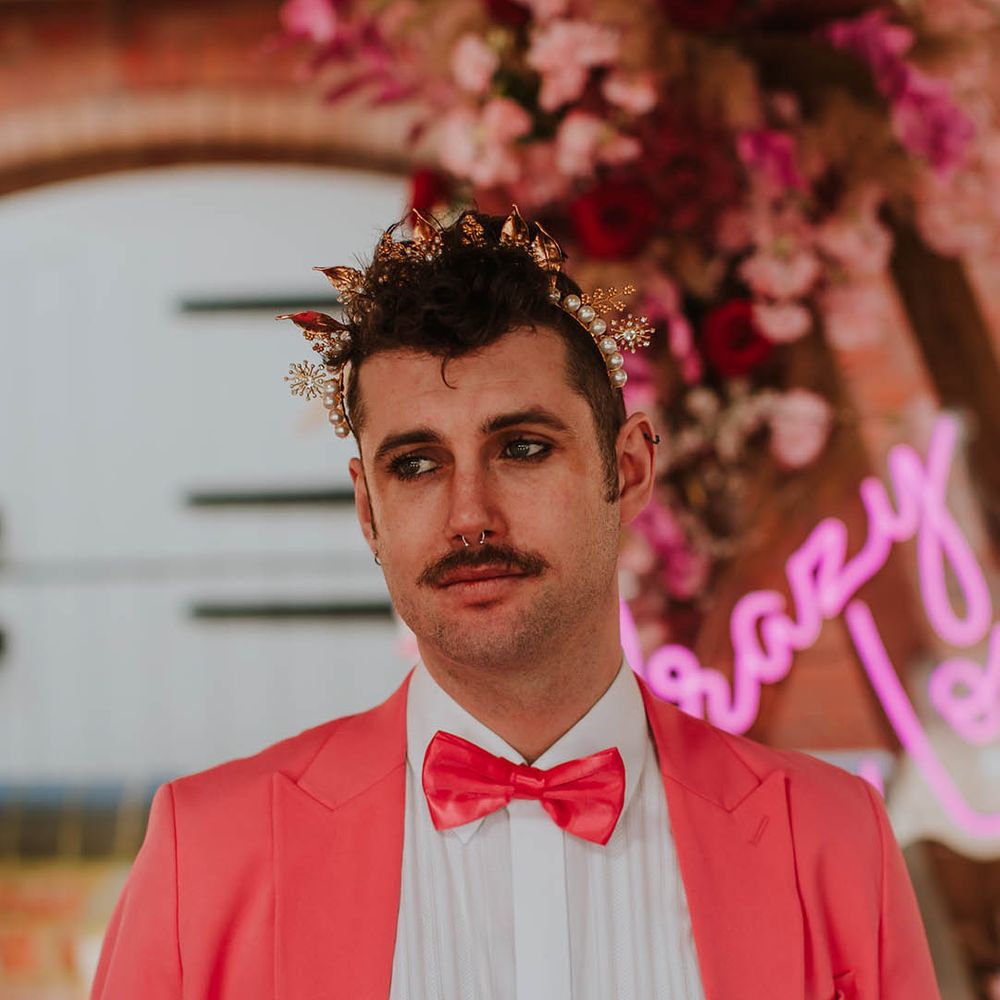Groom in a pink suit and satin bow tie wearing a gold crown headpiece 