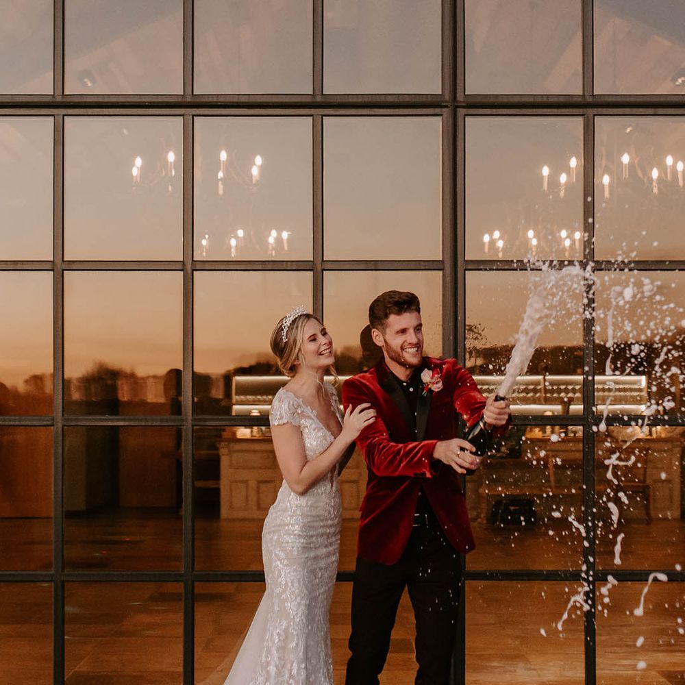 Bride in fitted lace wedding dress and pearl tiara, groom in red velvet tuxedo doing champagne pop