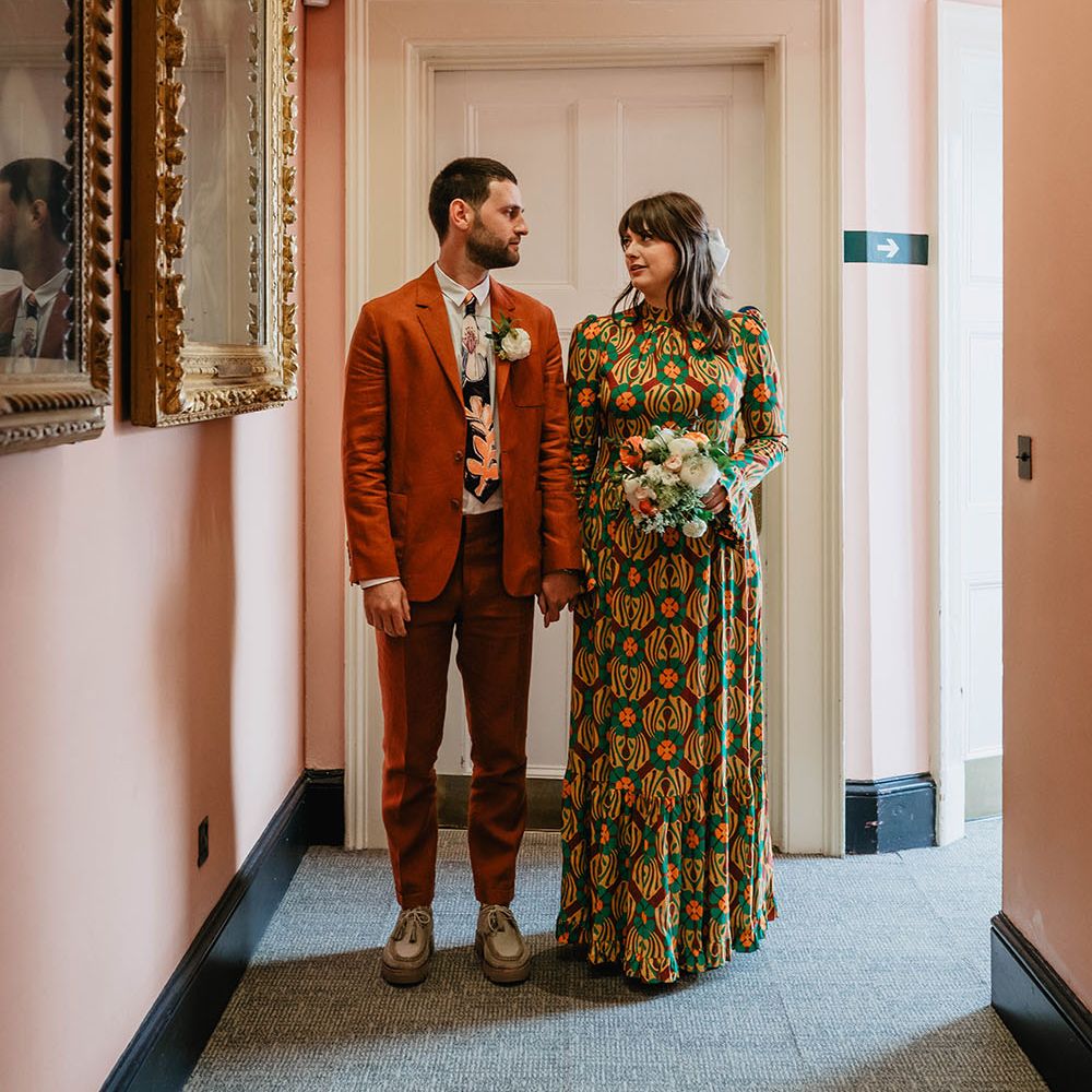 Bride & groom hold hands as groom wears orange suit and bride wears multicoloured gown and holds white and orange bridal bouquet 