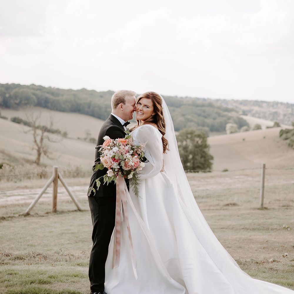 Bride holds colourful floral bouquet tied with silk pink ribbon stands with her groom outdoors 