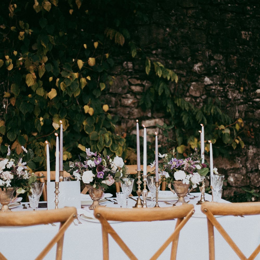 Wedding tablescape with lavender and white tapered candles in gold candle holders and mixed neutral flower arrangements with white carnations, purple tulips, roses and foliage 