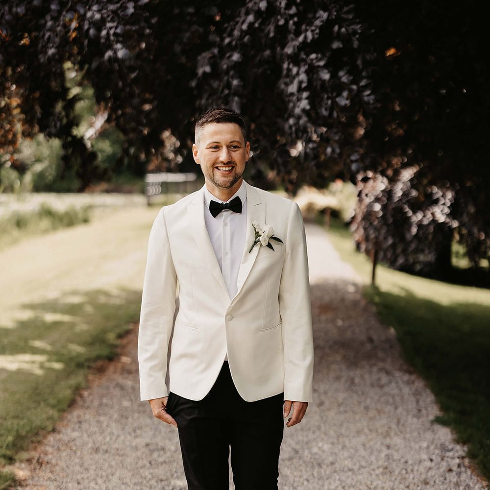 Groom in black suit trousers, white suit jacket, black bow tie, and white rose buttonhole 