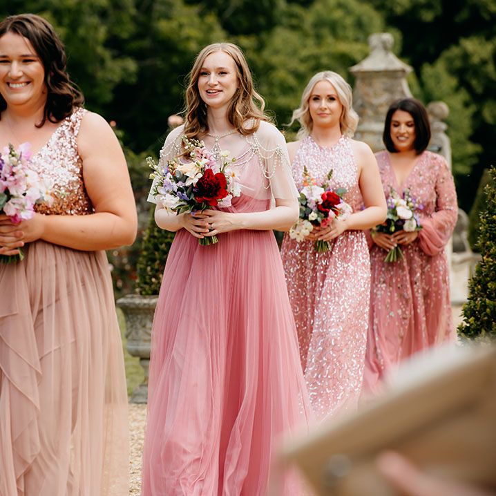 Bridesmaids in mismatched pink and sparkly sequin dresses holding colourful bouquets as they walk down the aisle of the outdoor ceremony 