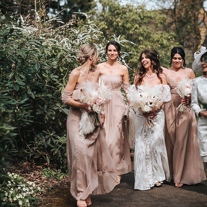 Bridesmaids in neutral pink dresses walking with the bride in her lace gown and the mother of the bride in a matching white dress and hat