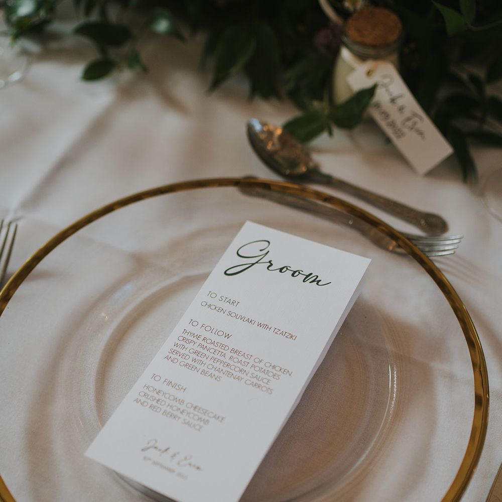 Black and white wedding menu on the clear charger plates with gold rim on white tablecloth with foliage table runner