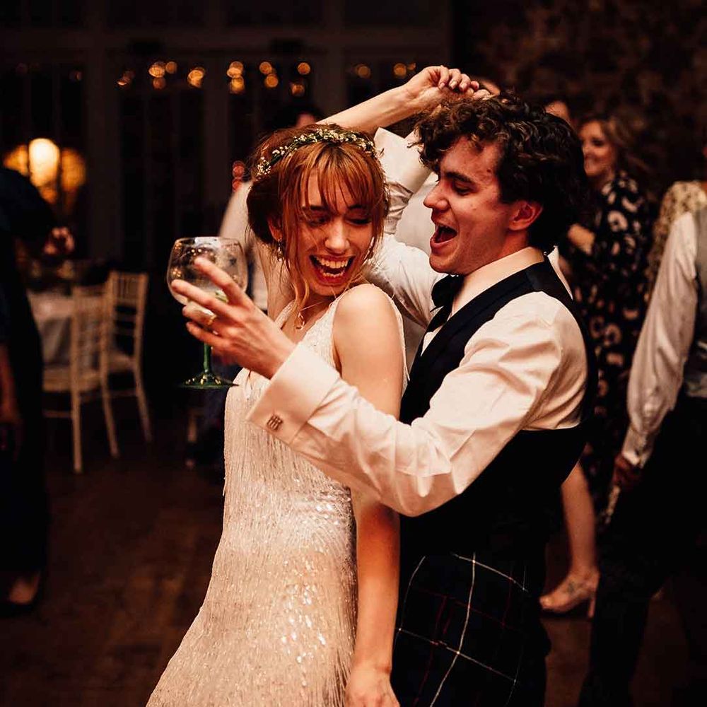 Bride in short fringed wedding dress and ethereal flower crown dancing with groom in waistcoat and white shirt at Oxnead Hall 